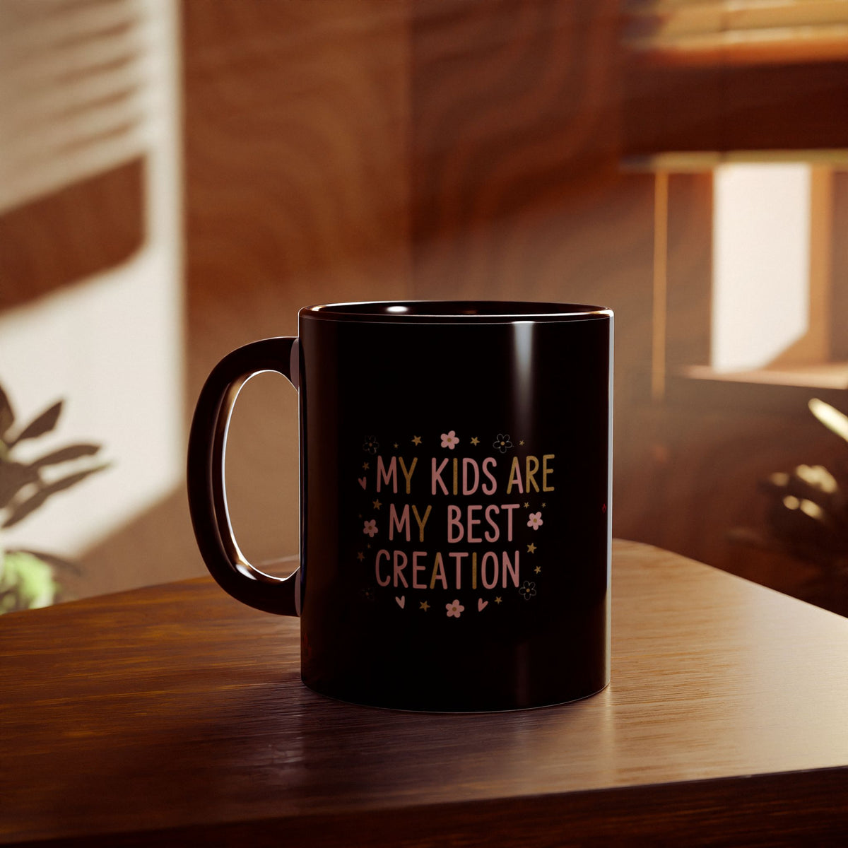 A black ceramic coffee mug with the text 'MY KIDS ARE MY BEST CREATION' in gold and pink lettering, decorated with small pink flowers and gold stars, sitting on a wooden table in a warm, sunlit room.