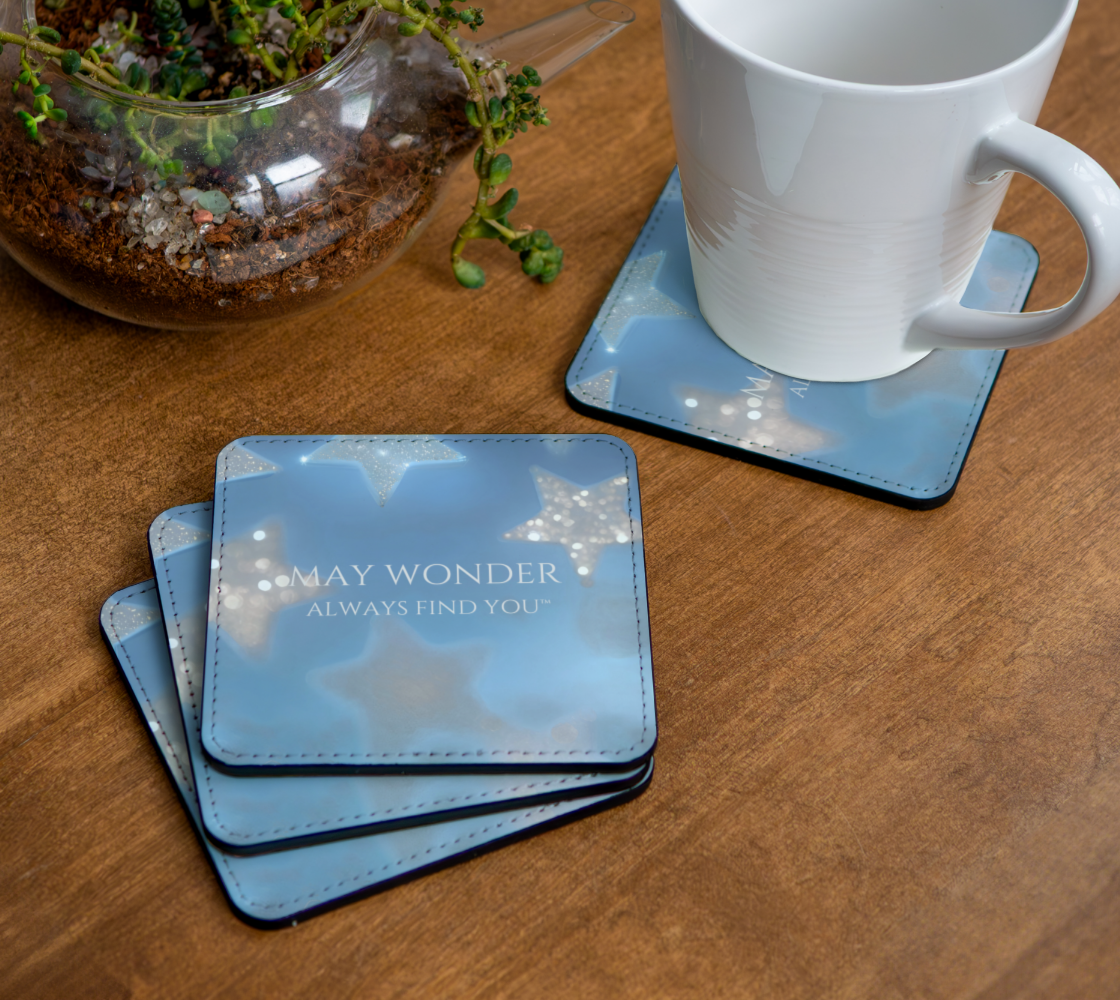 A stack of square light blue coasters with a starry night sky design and the text 'MAY WONDER ALWAYS FIND YOU' on a wooden table, alongside a white ceramic mug on a matching coaster and a glass terrarium.