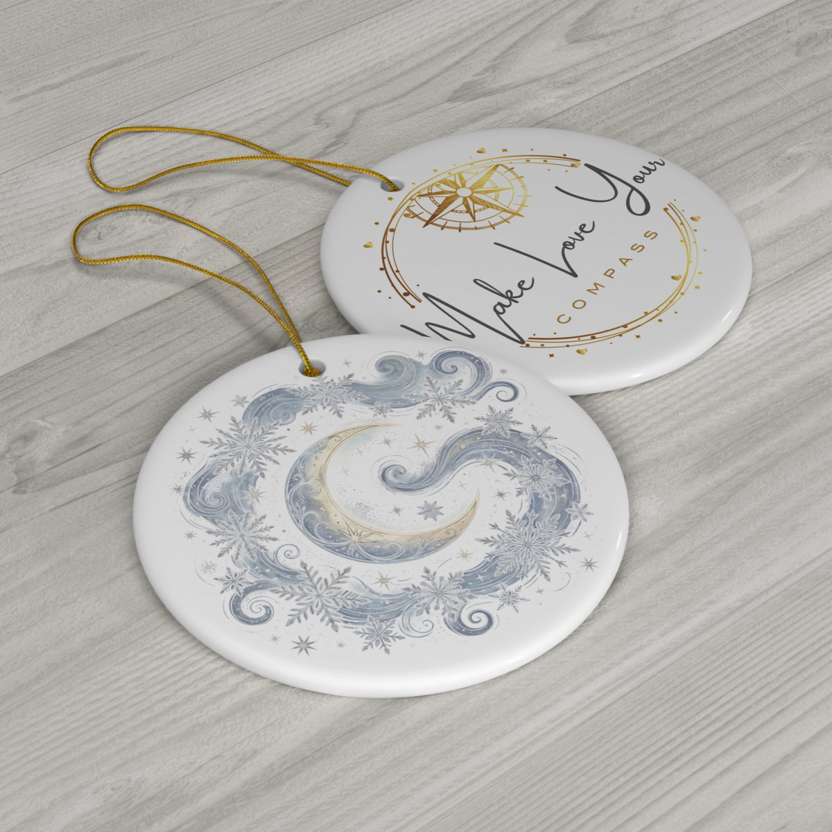 Two round white ceramic Christmas ornaments with gold hanging loops, displayed on a light gray wood background. The top ornament features a gold compass design with the words 'Make Your Compass' in black script.  The bottom ornament is of a moon and compass with snowflakes. 