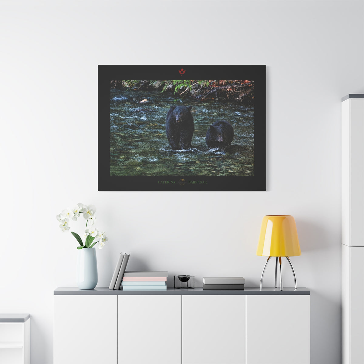A rectangular black-framed print or plaque featuring a photograph of two black bears standing in a shallow stream, with a small red maple leaf emblem at the top and the words 'CATERINA BARREGAR' at the bottom.