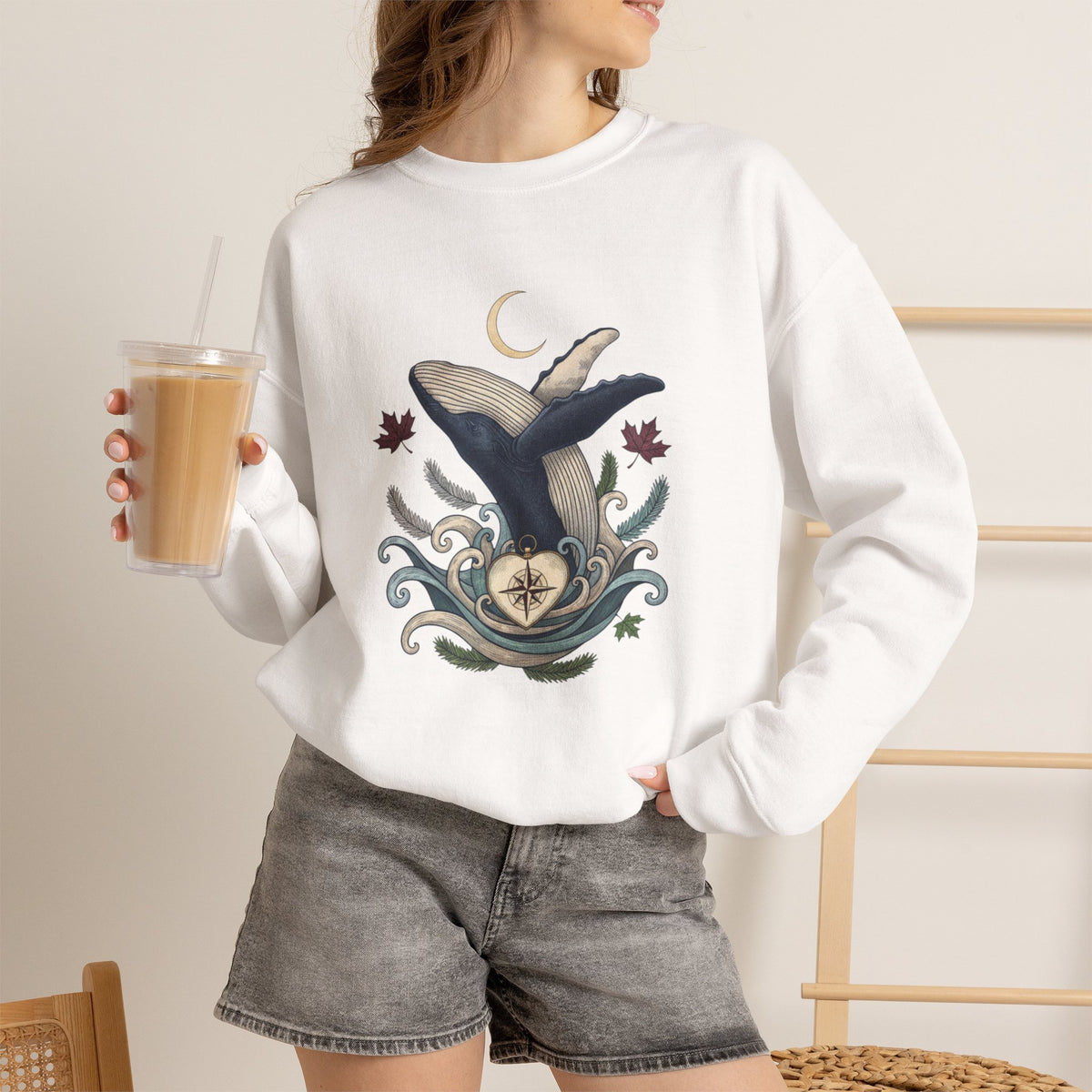 Make Love Your Compass Whale Moon Sweatshirt: A lifestyle photo of a woman wearing the white sweatshirt variant while holding an iced coffee tumbler in a cozy indoor room setting.