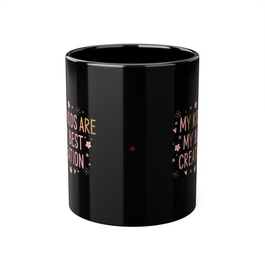 A glossy black ceramic mug with gold and pink text reading 'Kids are my greatest creation'  decorated with small pink flowers and gold stars, shown on a plain white background.

