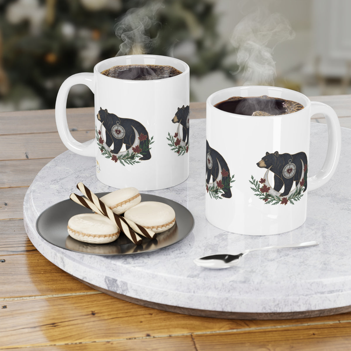 Two white ceramic mugs with black bear illustrations and a pine wreath with Canadian flag accents, filled with steaming coffee and displayed on a round marble tray with macarons and a spoon on a wooden table.