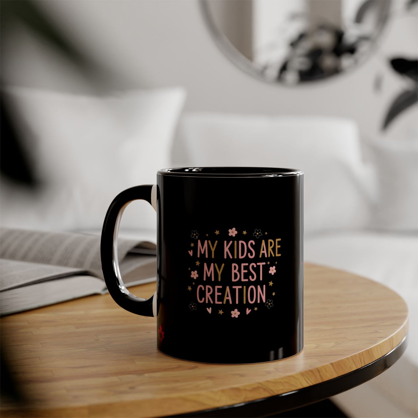 A black ceramic mug with the text 'MY KIDS ARE MY BEST CREATION' in pink and yellow, sitting on a wooden table in a cozy, softly blurred living room setting.