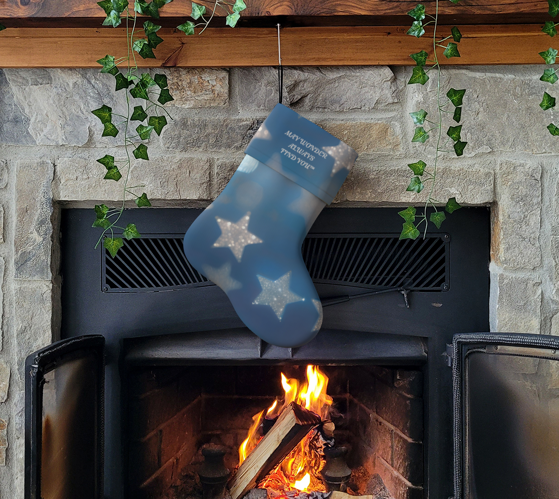 A blue Christmas stocking with white star patterns hangs from a fireplace mantel above an active wood-burning stove. The stocking has white text reading 'May Wonder Always Find You' and is set against a stone fireplace with green ivy vines.