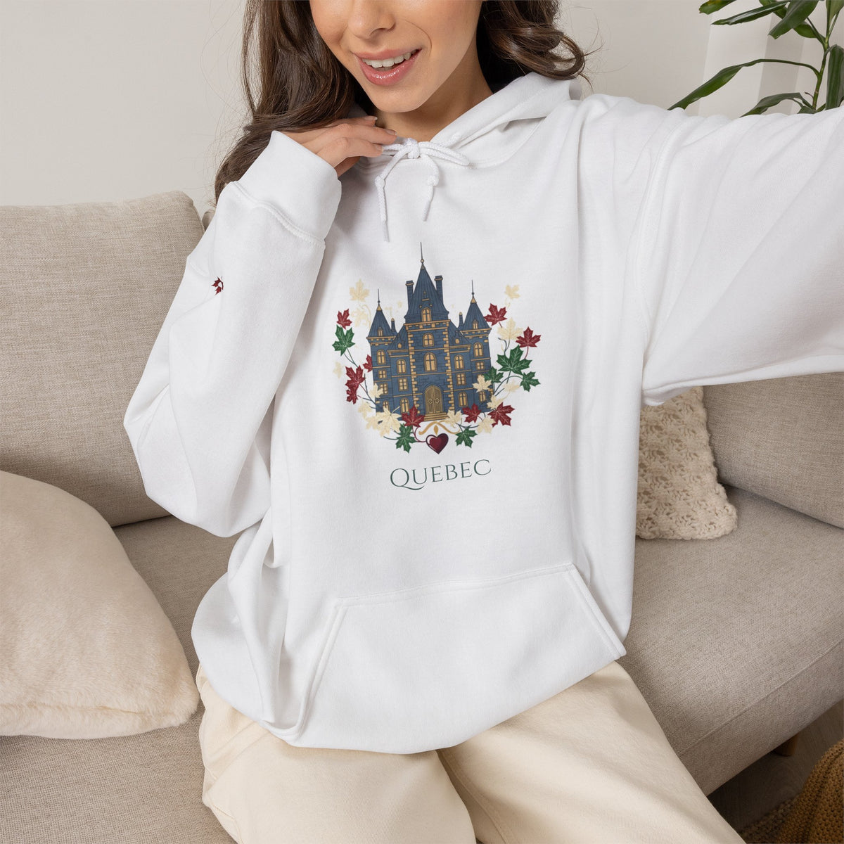 A woman wearing a white pullover hoodie with a colorful graphic of a castle and floral design, with the word 'QUEBEC' printed below, sitting on a beige couch.