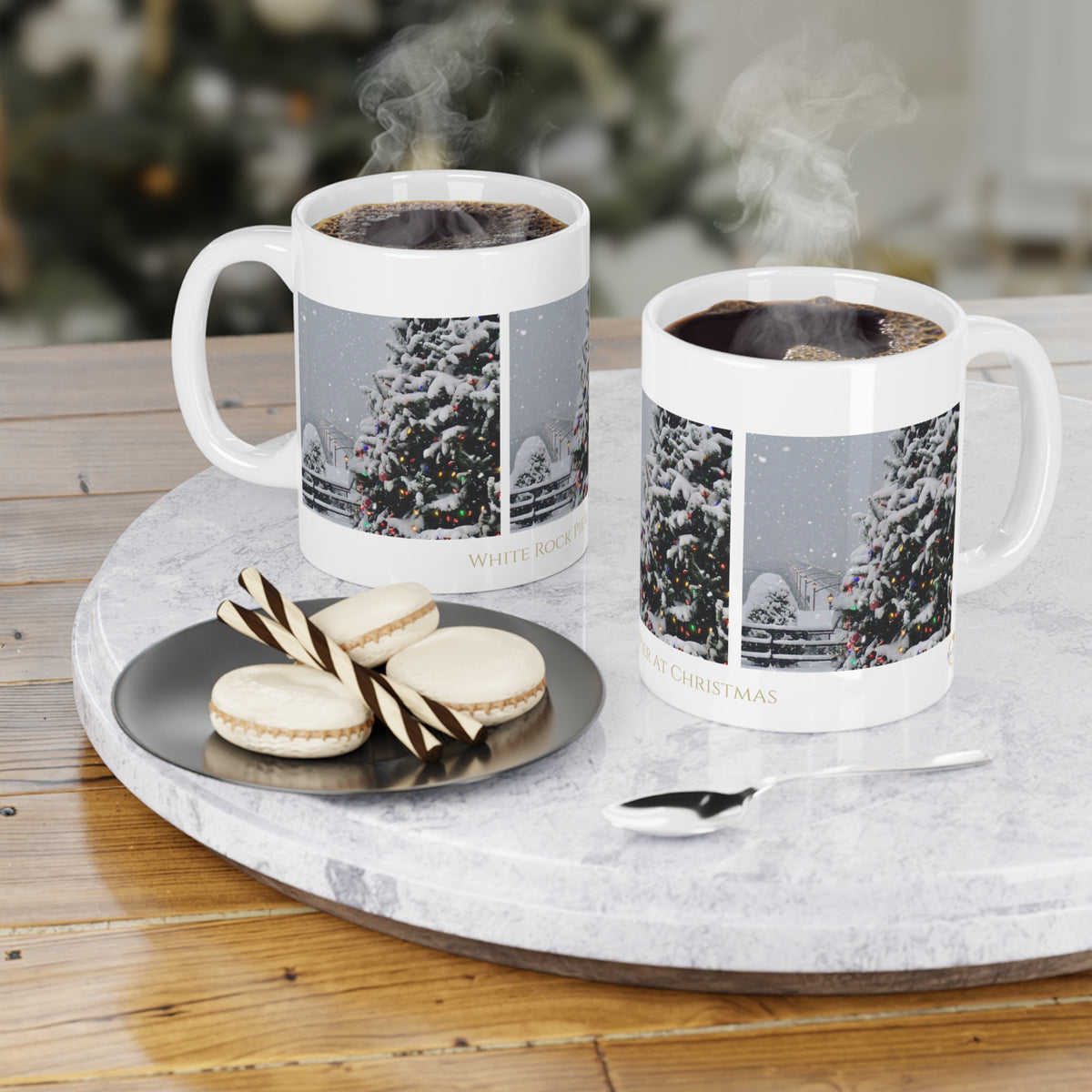Two white ceramic mugs filled with hot coffee, printed with a winter Christmas tree scene and the words 'White Rock Pier At Christmas' displayed on a round marble tray with macarons and a spoon.